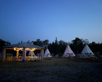 An outdoor scene at dusk with a stone pavilion and several tipis arranged in a circle with bleachers.
