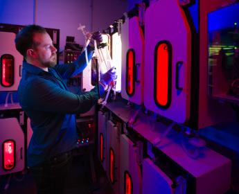 A scientist inside a lab pulls a large, clear cylinder from inside one of several illuminated chambers.