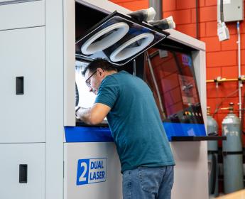 A man leans into the building chamber of a large 3D printer. A control panel is to his right. 