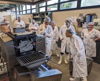 Seven people wearing boots, disposable hairnets, and white lab coats, along with protective goggles, are gathered around a MilkyLab machine in a laboratory located in a basement bathed in light from a row of windows at the very top and all along the wall. Some of them are looking at their cell phones, others are smiling, and still others have approached the cheese-making machine and seem to be wondering how it works.