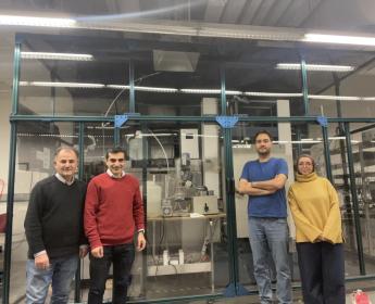 Trois hommes et une femme se tiennent devant une grande section vitrée d’un laboratoire rempli de matériel expérimental.
