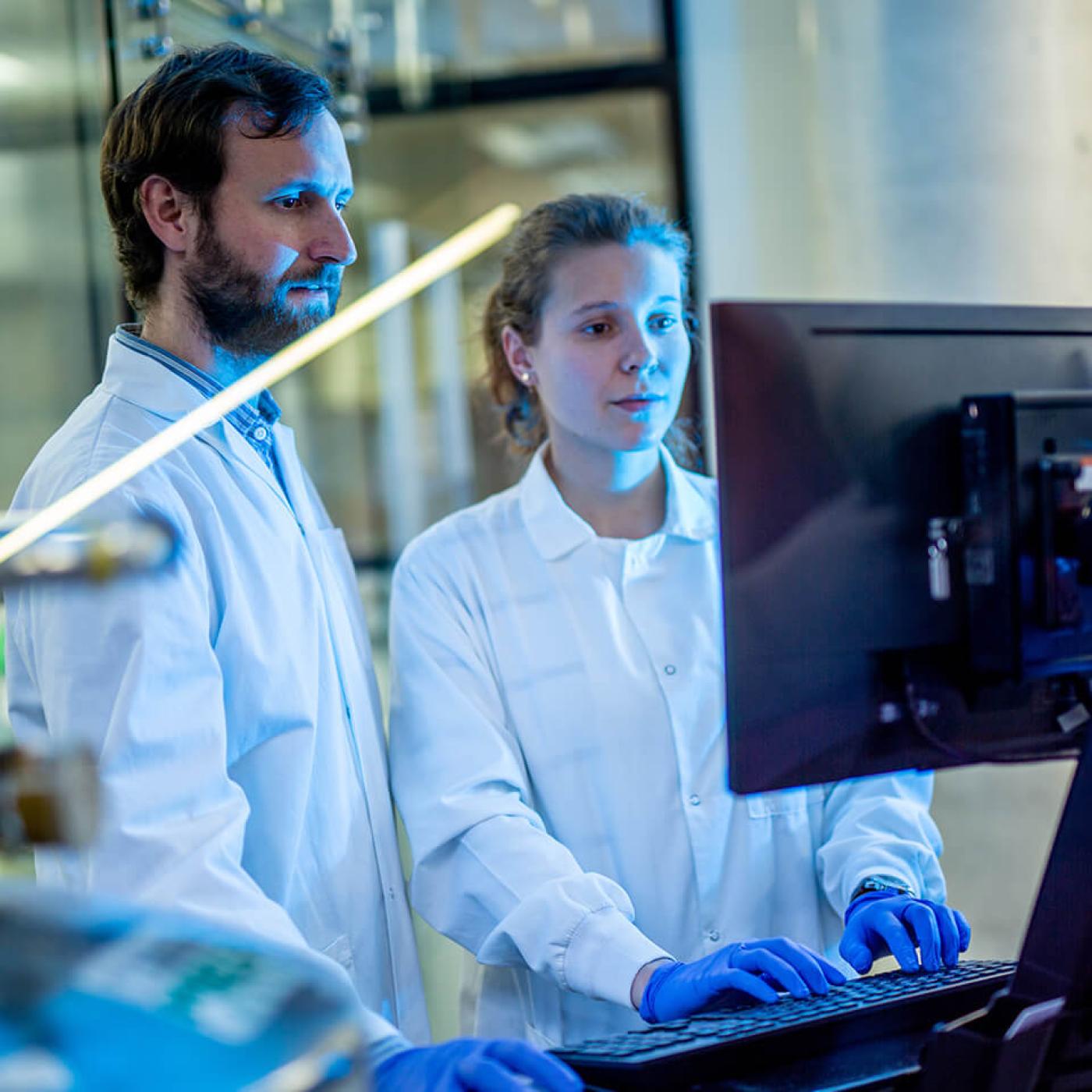 Two people wearing lab coats and gloves look at a computer screen