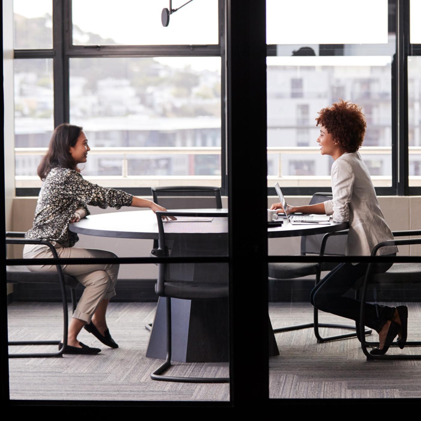 Sideview of two women sitting at a meeting table in an office in front of windows