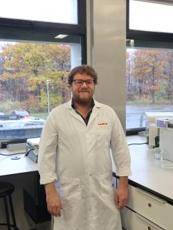 Photo of a smiling man standing in front of windows, shot from the head down to the thighs, wearing a white lab coat. He has slightly reddish hair, a beard, and a mustache. The background features autumn trees, yellow and orange, reflected in puddles of rainwater. 