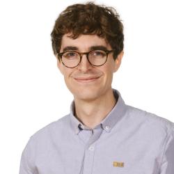 A portrait of a man with brown hair, thick eyebrows, and glasses, smiling broadly. He is wearing a lilac-colored shirt.