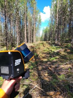 A hand points an orange device roughly the size of an electric drill at a group of trees.