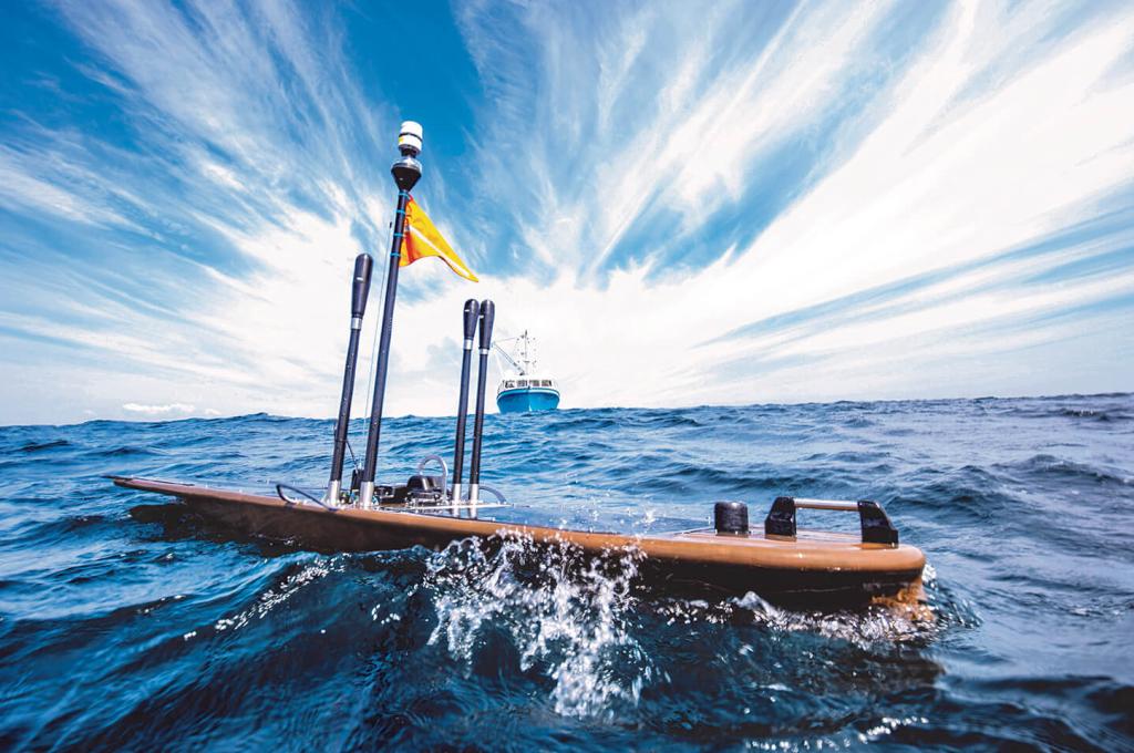 A vessel the size and shape of a surfboard with an apparatus attached glides on the surface of the ocean with a ship in the background.