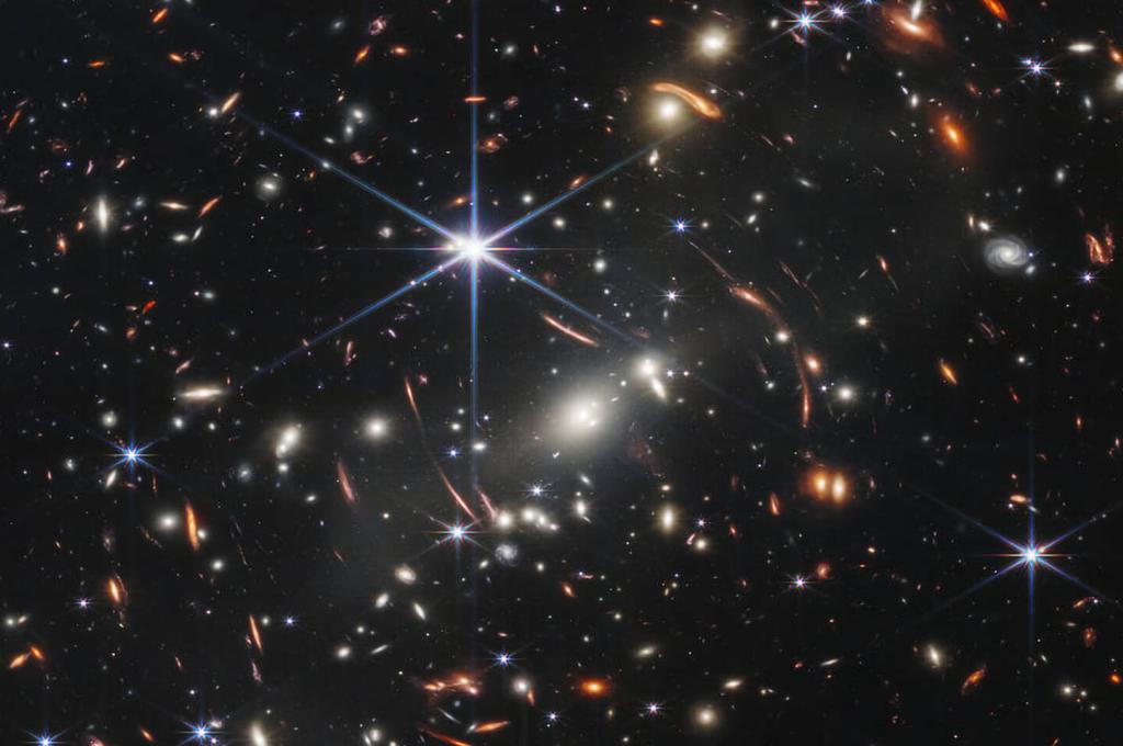 Des étoiles au premier plan apparaissent brillantes avec des pointes de diffraction à six points, en raison de la forme des miroirs du télescope James-Webb. Il s’agit de l’amas de galaxies SMACS 0723, qui agit comme une lentille gravitationnelle, déviant la lumière des galaxies plus lointaines, qui apparaissent comme des taches lumineuses plus brouillées.
