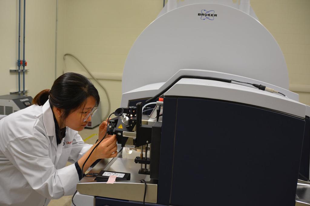 A person in a lab coat looks closely at an apparatus that is part of a black and grey machine in a lab.