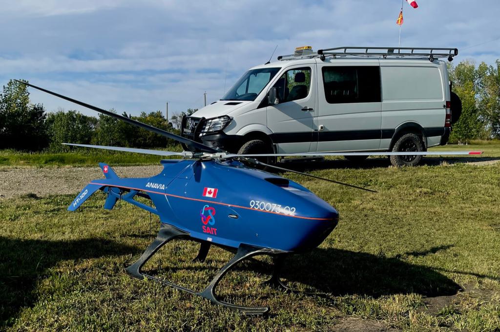 Un drone ressemblant à un petit hélicoptère bleu est posé sur une zone herbeuse au premier plan, tandis qu'une camionnette blanche est garée dans une allée à l'arrière-plan.