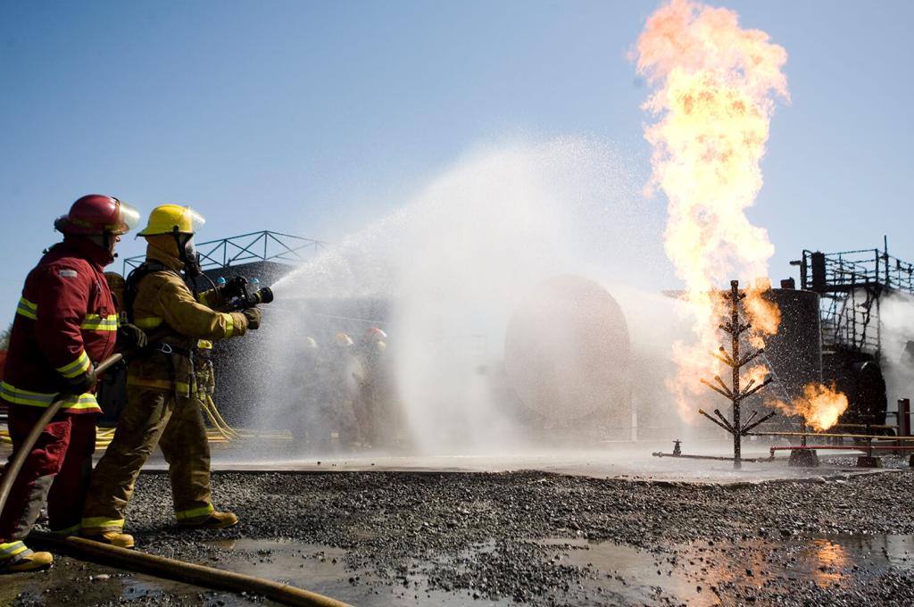 Photo prise lors d’un exercice de simulation d’un incendie. Deux personnes en habit de combat de feu dirigent une lance à incendie vers un bâtiment en flamme