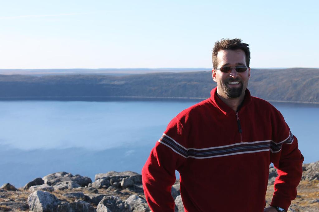 Guillaume St-Onge is wearing a striped red sweater and sunglasses, posing in front of the Pingualuit crater in Quebec, full of clear blue water.