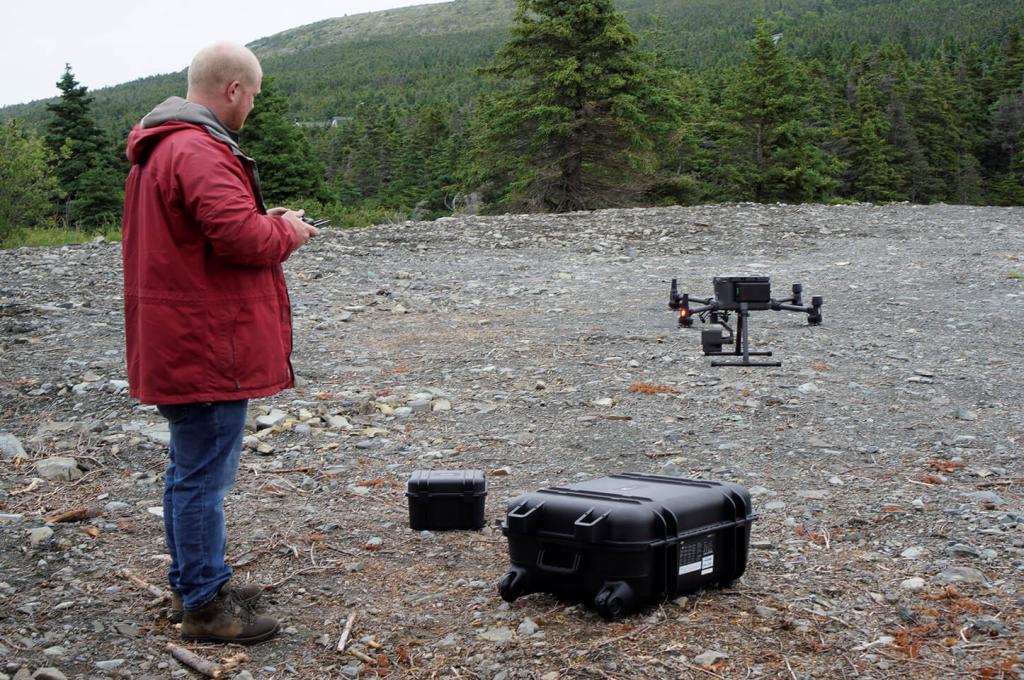 A man in a red jacket holds the remote control as a drone takes off from a rubble-strewn site. 