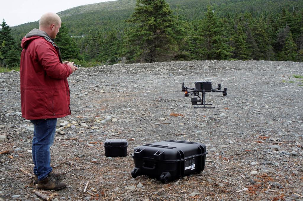 Un homme vêtu d’un manteau rouge tient une télécommande alors qu’un drone décolle d’un site jonché de débris.