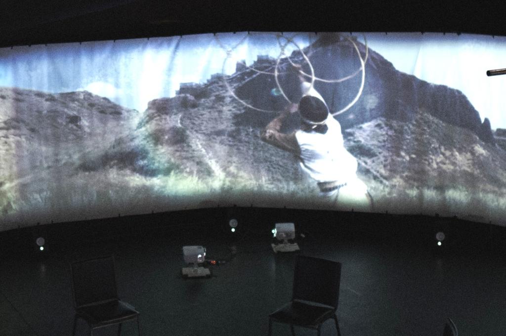 A person hoop dances on a panoramic screen in a large circular space.
