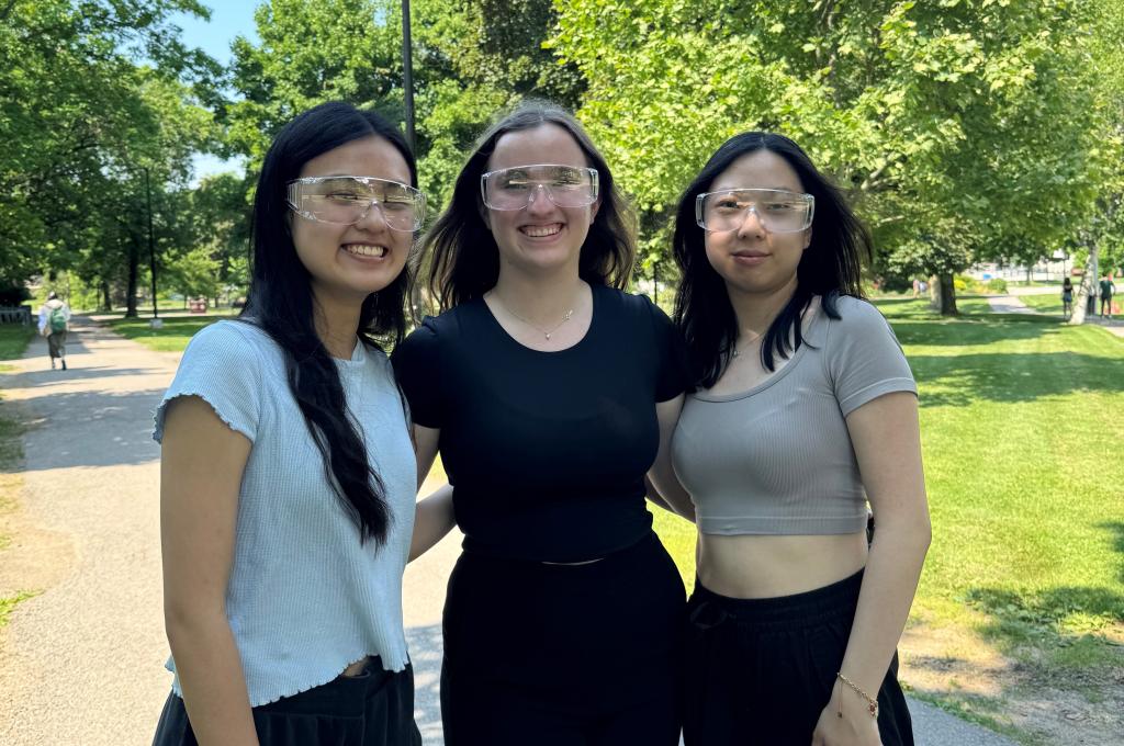 Three young women pose in an outdoor setting wearing clear plastic safety-style glasses