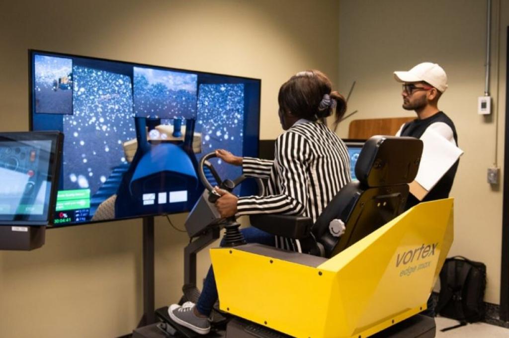 Vue de dos d’une personne assise aux commandes d’un simulateur dont elle tient le volant sous le regard d’une autre personne à sa droite qui porte une casquette blanche.