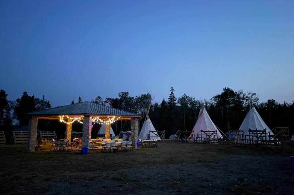 An outdoor scene at dusk with a stone pavilion and several tipis arranged in a circle with bleachers.