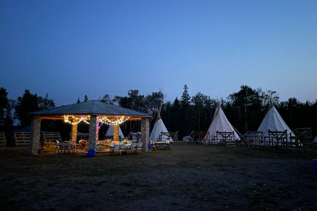 An outdoor scene at dusk with a stone pavilion and several tipis arranged in a circle with bleachers.