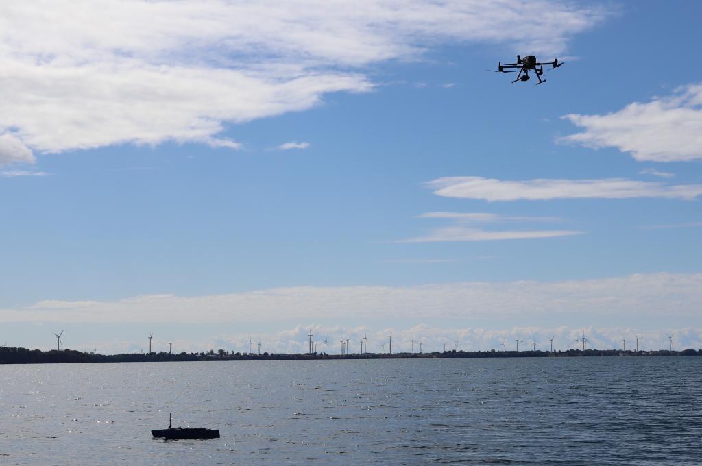 A quadrotor UAV flies in the upper right of the photo; in the water below floats a small uncrewed vessel Greef and her team plan to use as a recharging station. 