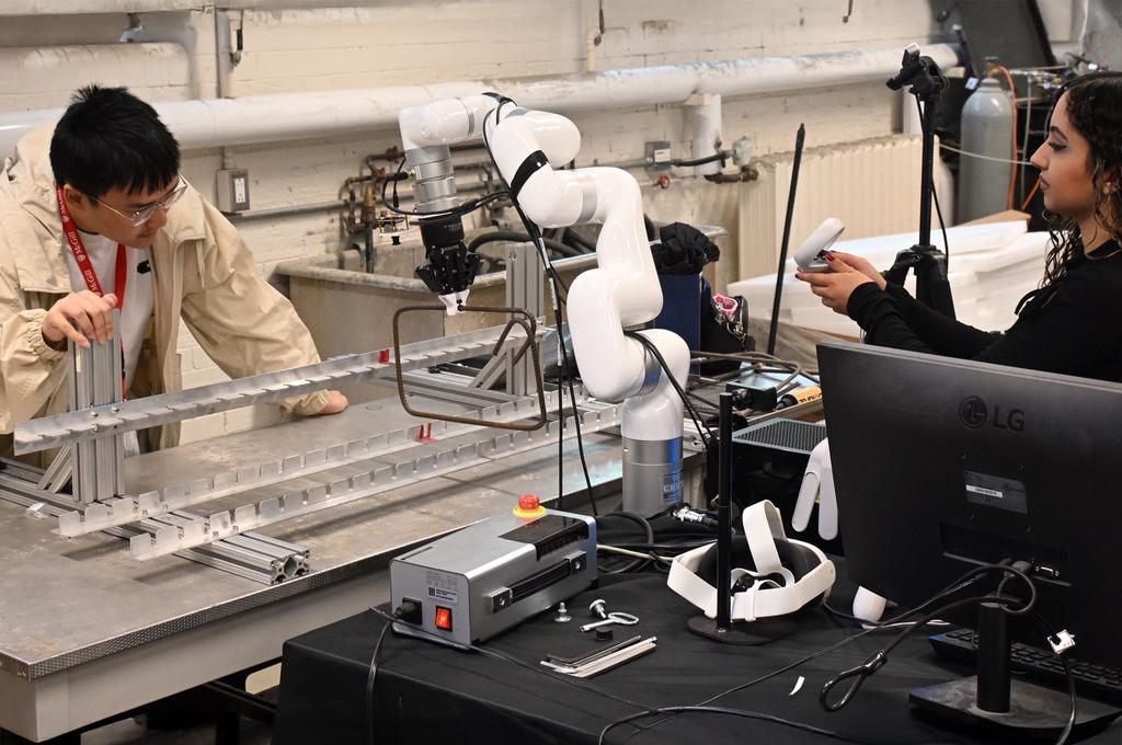 A multi-hinged robotic arm holds a rectangle formed from rebar. On the right, a young woman holds a small white box in her hands. On the left, a young man looks at the frame into which the rebar is being inserted.