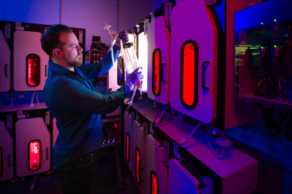 A scientist inside a lab pulls a large, clear cylinder from inside one of several illuminated chambers.