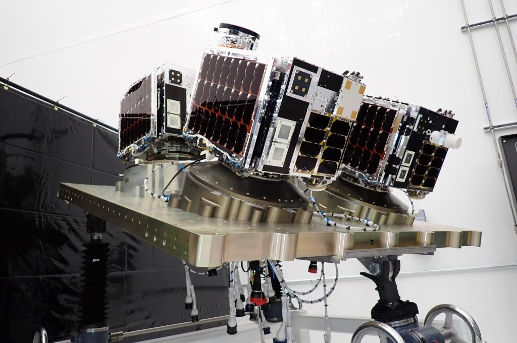 Three box-shaped satellites mounted on a metallic platform in a lab.