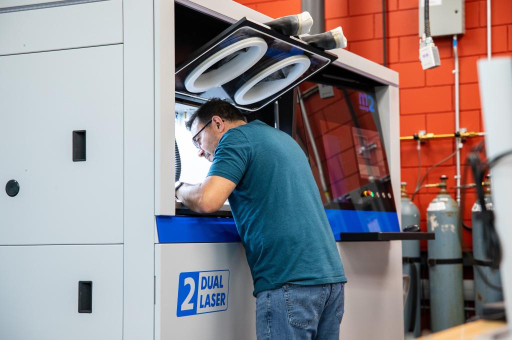 A man leans into the building chamber of a large 3D printer. A control panel is to his right. 