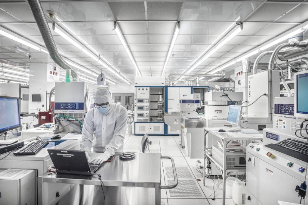 A researcher in hooded white cleanroom coveralls, mask and gloves types on a laptop in a white-tiled cleanroom packed with high-tech equipment.