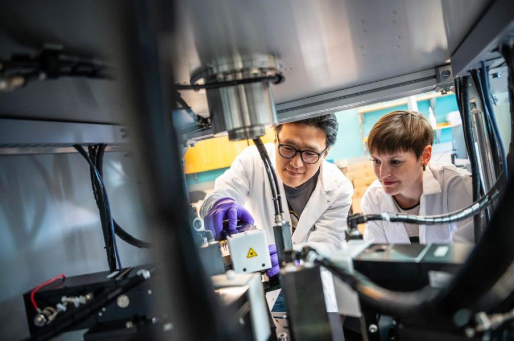 Two scientists in lab coats and gloves examine the contents of a metal chamber equipped with high-tech components.