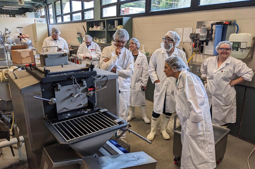 Seven people wearing boots, disposable hairnets, and white lab coats, along with protective goggles, are gathered around a MilkyLab machine in a laboratory located in a basement bathed in light from a row of windows at the very top and all along the wall. Some of them are looking at their cell phones, others are smiling, and still others have approached the cheese-making machine and seem to be wondering how it works.