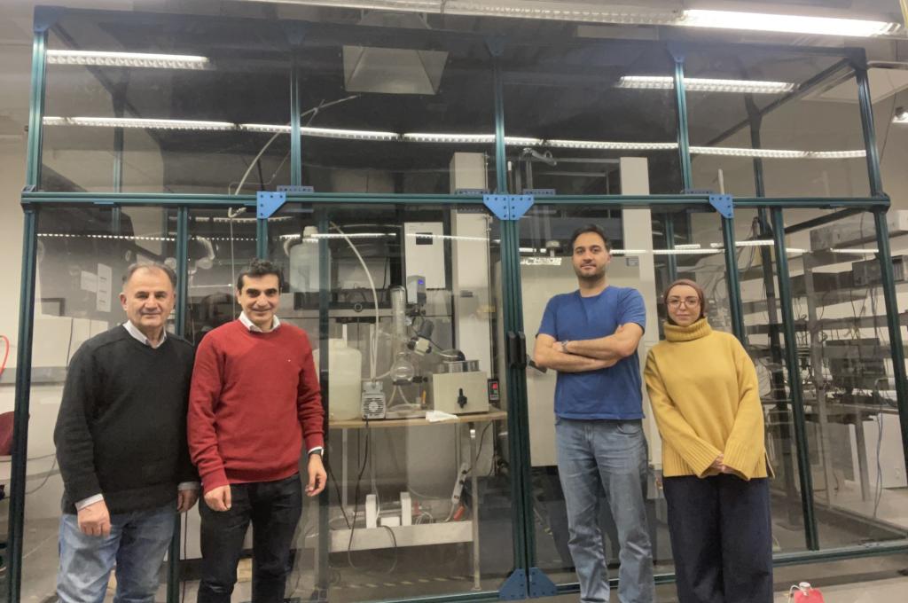 Four researchers stand in front of a large, glassed-off section of a lab filled with experimental equipment.