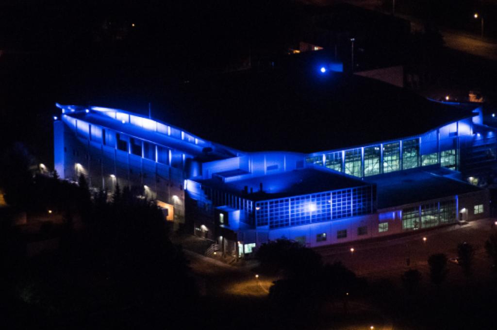 A large building seen from above at night. The sides are lit up with bright blue light.