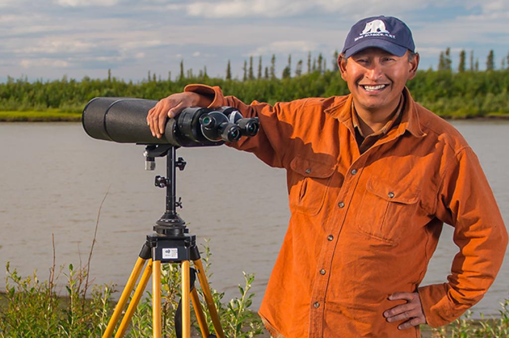 A man stands beside a river with his arm resting on a set of long-range observation binoculars.