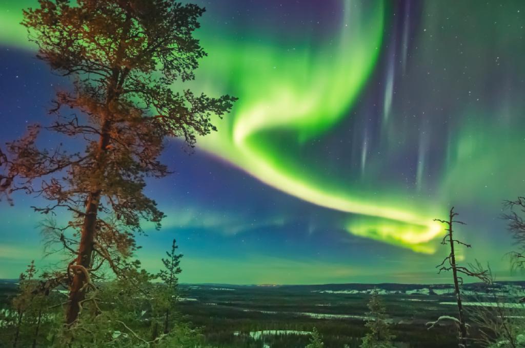 Un tourbillon de lumière verte dans le ciel au-dessus d’un paysage peu boisé.