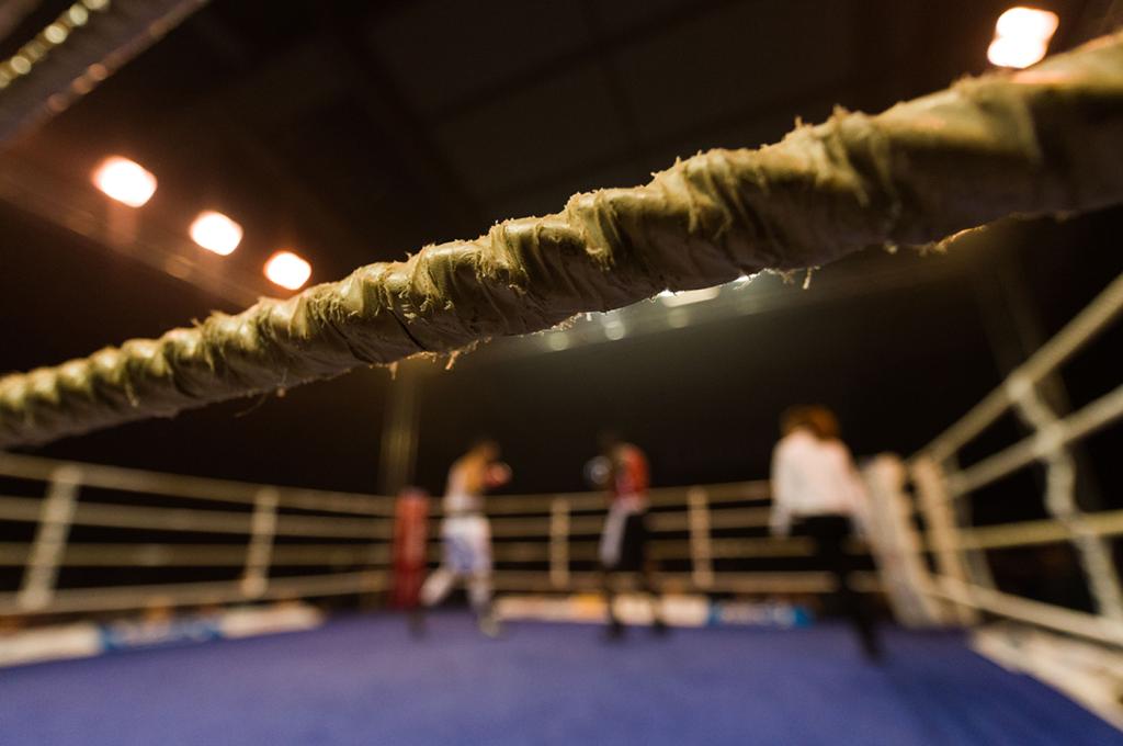 Looking through the ropes into a boxing ring, with boxers blurred in the background.