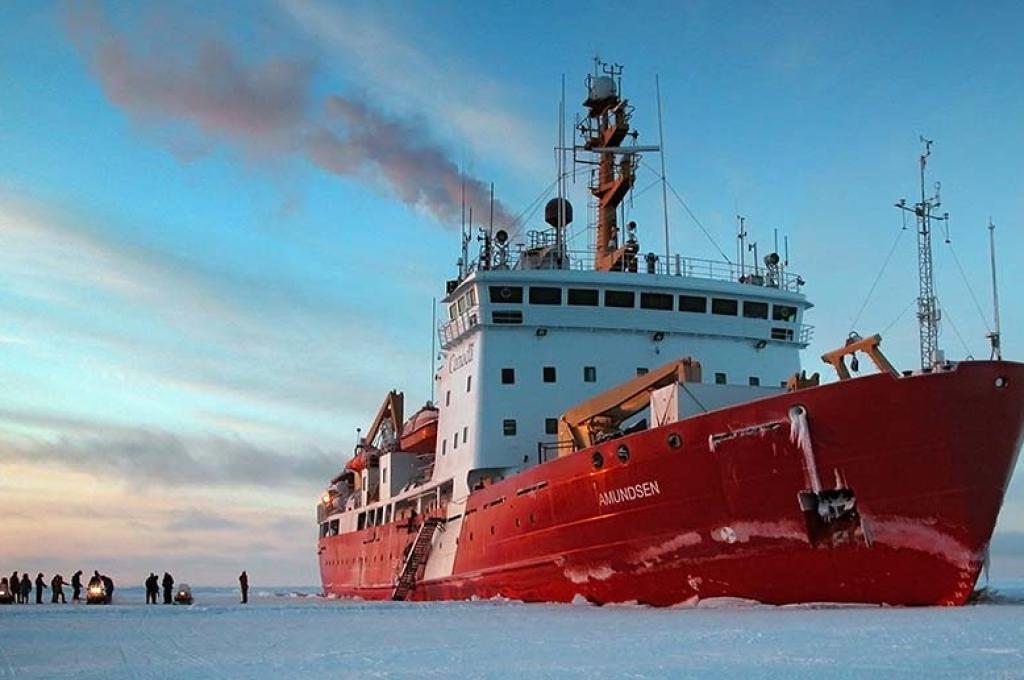 A small group of people stand on a vast expanse of ice, dwarfed by the red and white ice breaker beside them. 