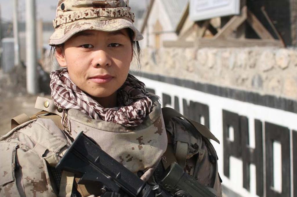 A woman in a military uniform faces the camera, angling a machine gun at the ground.