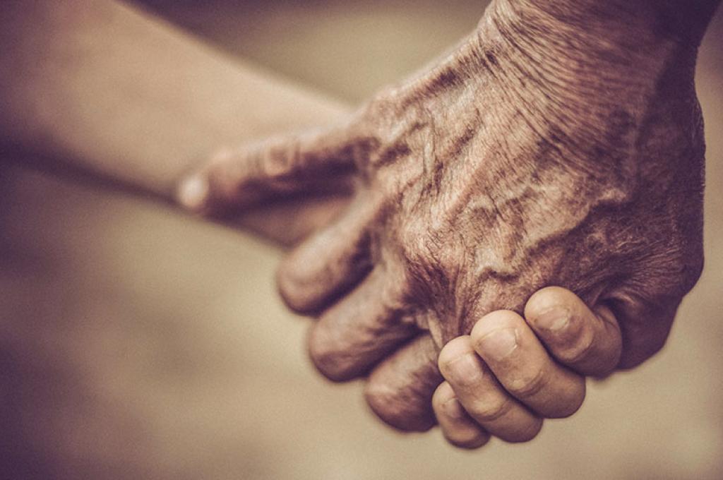 A close up of two hands joined against a blurred out, grey-brown background. The hands are wrinkled and aging.