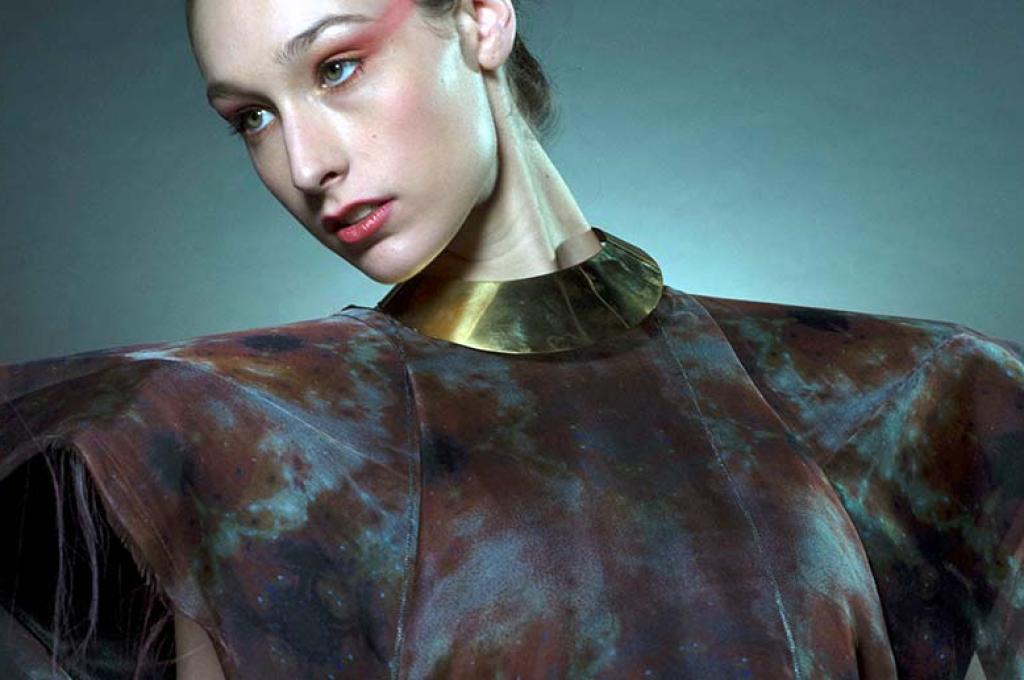 A woman wearing dramatic red eye shadow poses in a futuristic garment that can change shape or colour. The dress has high, pointed shoulders and dark brown and blue patterned material.