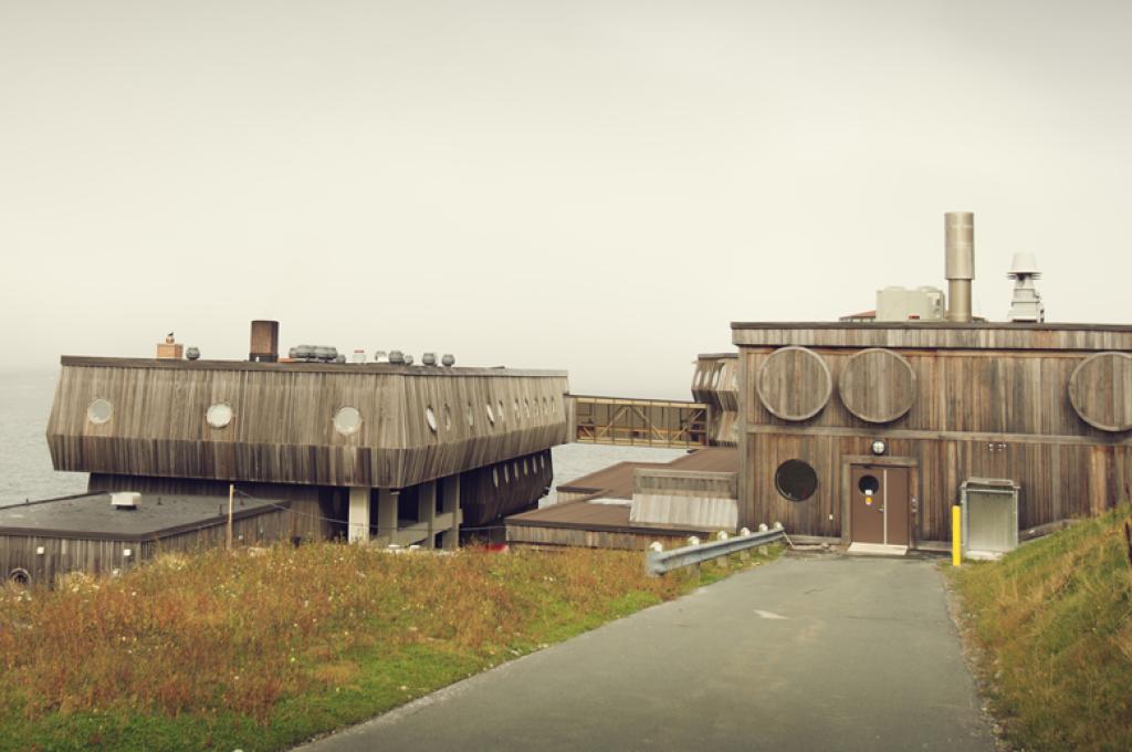 A wooden building resembling a ship with round portholes is perched on a coast line with fog on the opposite shore.