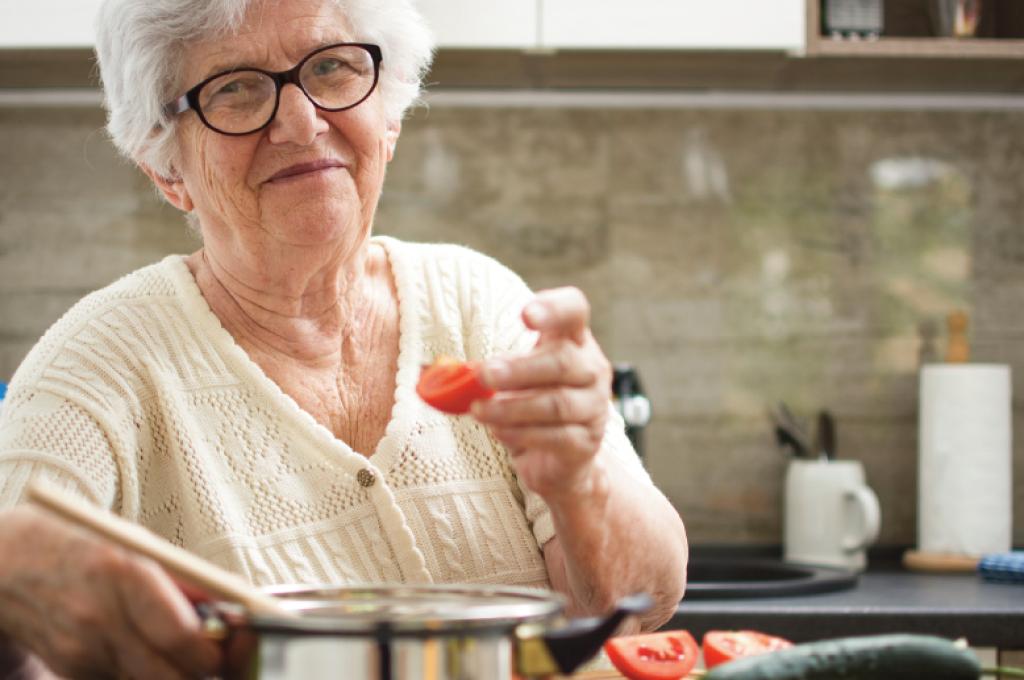 Un quartier de tomate à la main, une femme aux cheveux blancs se tient debout devant un chaudron sur une cuisinière