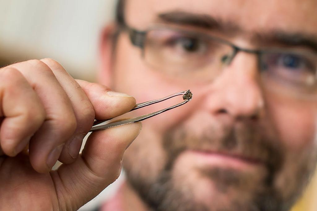 A man uses tweezers to hold up a small diamond. The diamond is in focus, blurring out the man’s face.