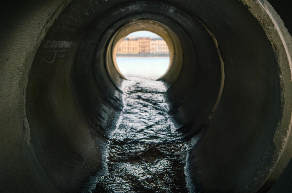 Un liquide noir s’écoule dans un tuyau en béton dont l’extrémité laisse entrevoir une rivière devant un paysage urbain