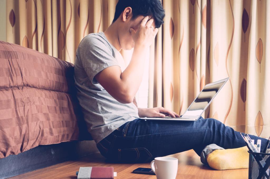 Une main posée sur le front, une personne assise par terre se penche sur un ordinateur portable