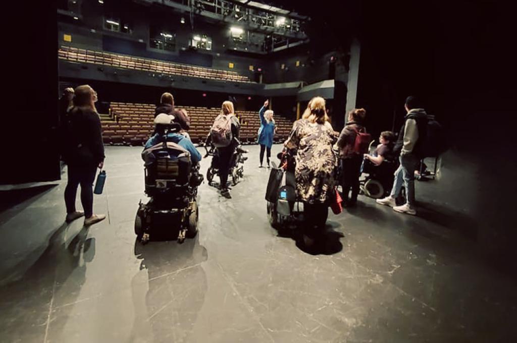 Depuis les coulisses, un groupe de personnes se tient sur une scène et regarde vers un auditorium vide.