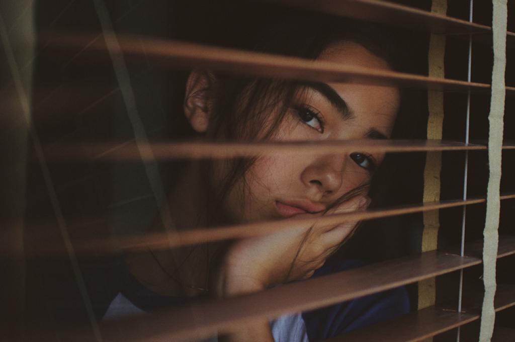 Une jeune femme regarde par la fenêtre à travers les volets ouverts, l’air ennuyé