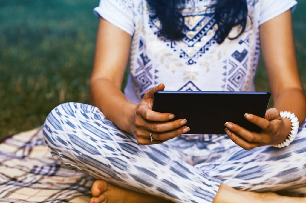 A woman sits cross-legged in the grass, holding an electronic tablet.