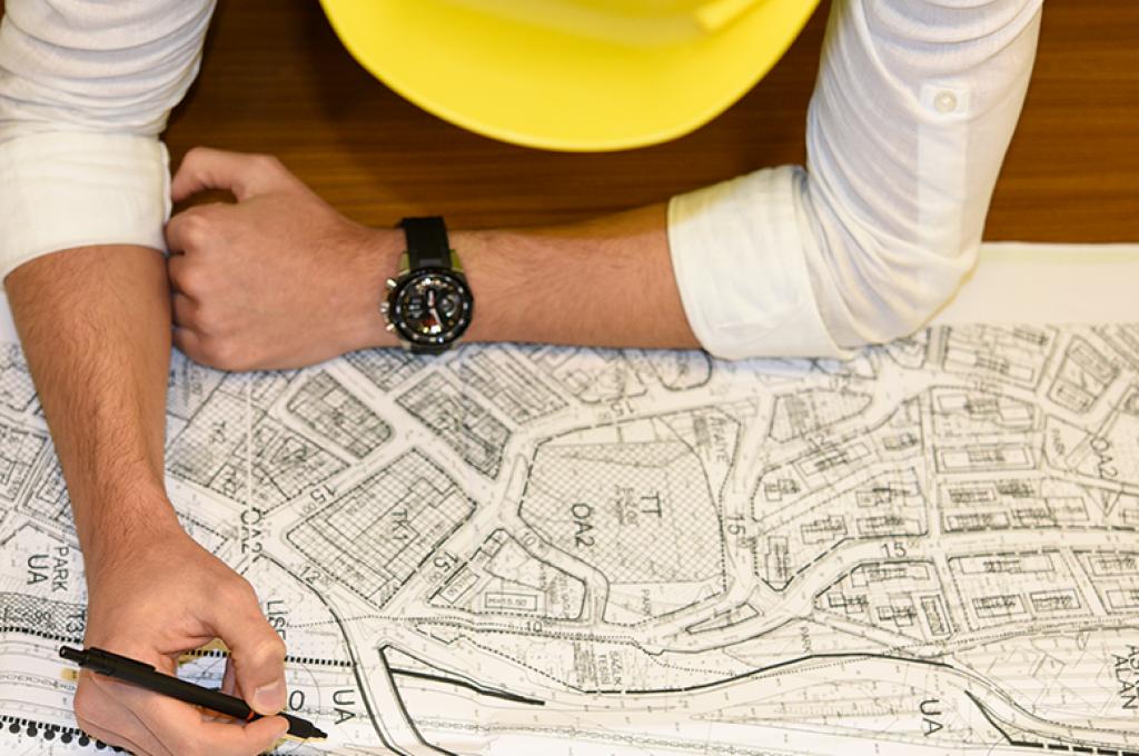 Vue du dessus d'une personne portant un chapeau de chantier jaune est penchée sur un plan de bâtiments posé sur une table en bois.