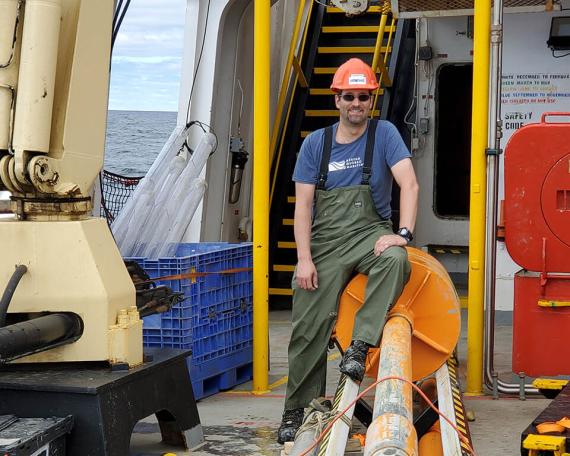 Guillaume St-Onge is wearing an orange hard hat, sunglasses and a fishing bib, seated on top of an orange core drill that is several metres long.